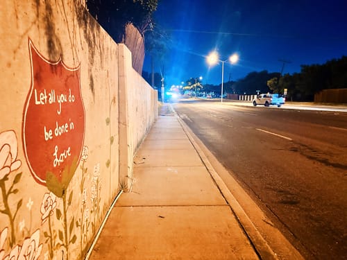 Wall, sidewalk, and road with a mural painted on the wall. "Let all you do be done in Love"